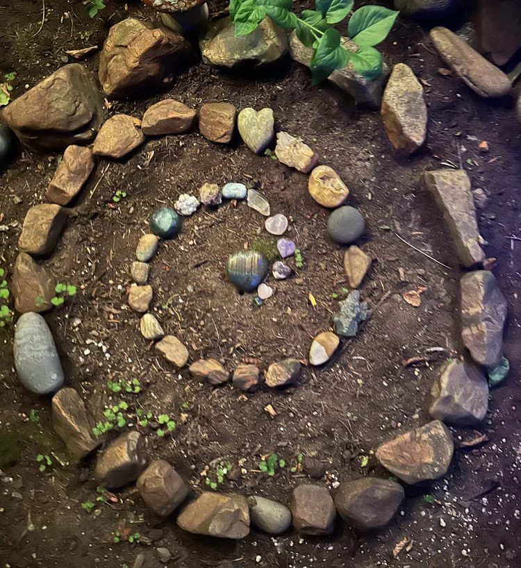 A spiral of stones laid by hand into the earth, a heart stone and a labradorite at its center, clover coming up between them.